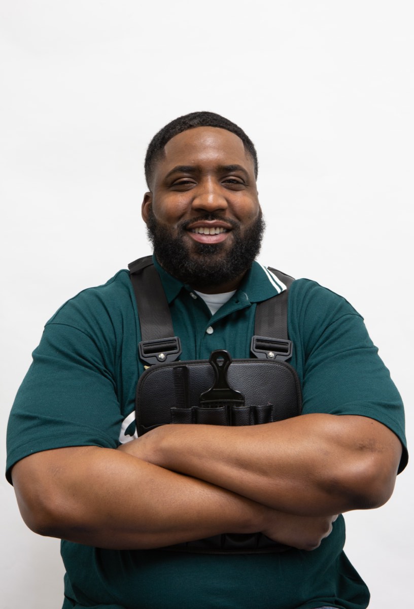 Portrait of a man in a green polo shirt with a stylist’s tool harness, against a white backdrop