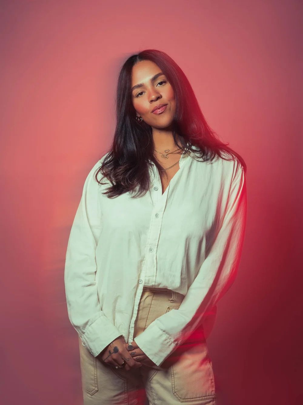 Portrait of a woman with long dark hair in a cream shirt and tan trousers, against a pink backdrop washed with red light