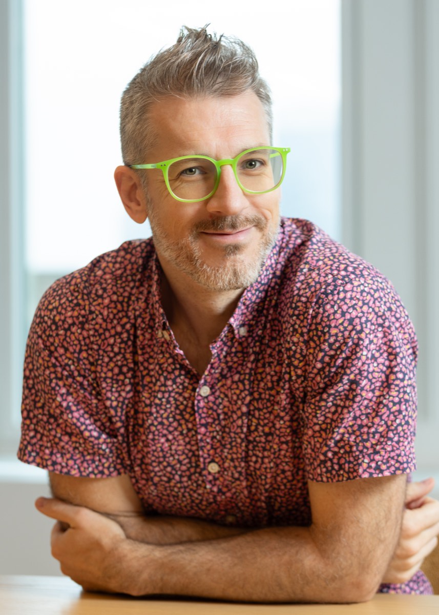Portrait of a man with green glasses and a pink patterned shirt, arms folded at a table