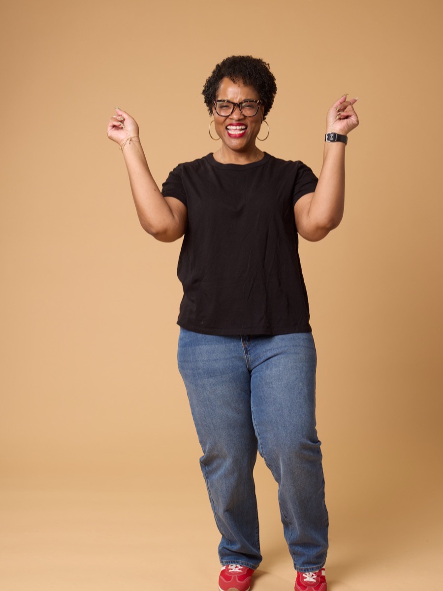 Portrait of a woman in a black t-shirt and jeans with both arms raised, against a warm tan backdrop