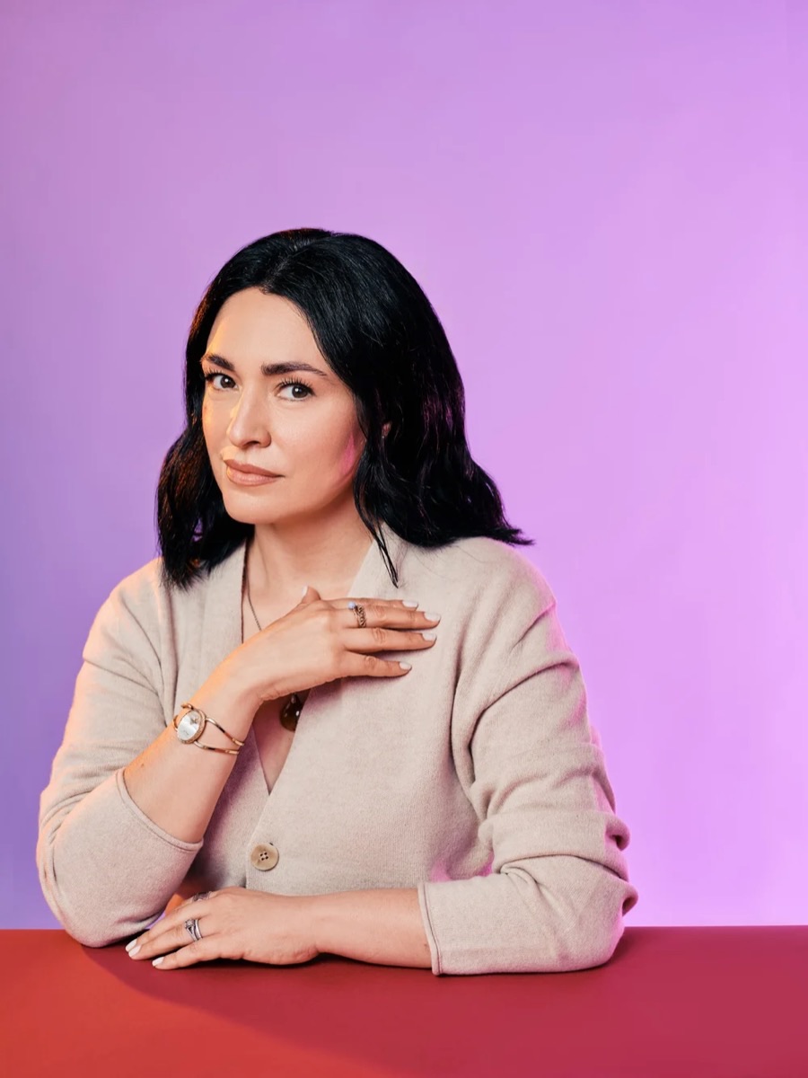 Portrait of a woman in a cream sweater seated at a red table, against a lilac backdrop