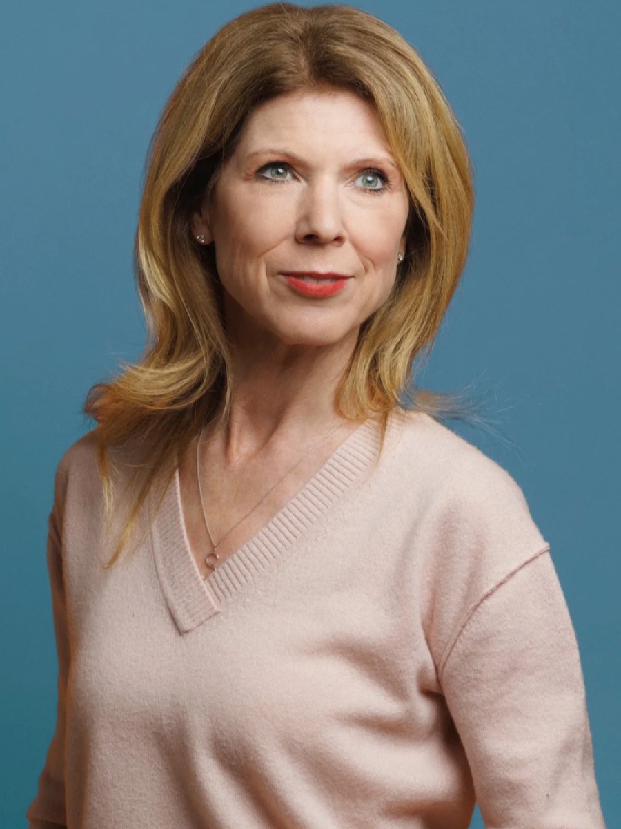 Portrait of a woman with blonde hair in a pale pink v-neck, against a blue backdrop