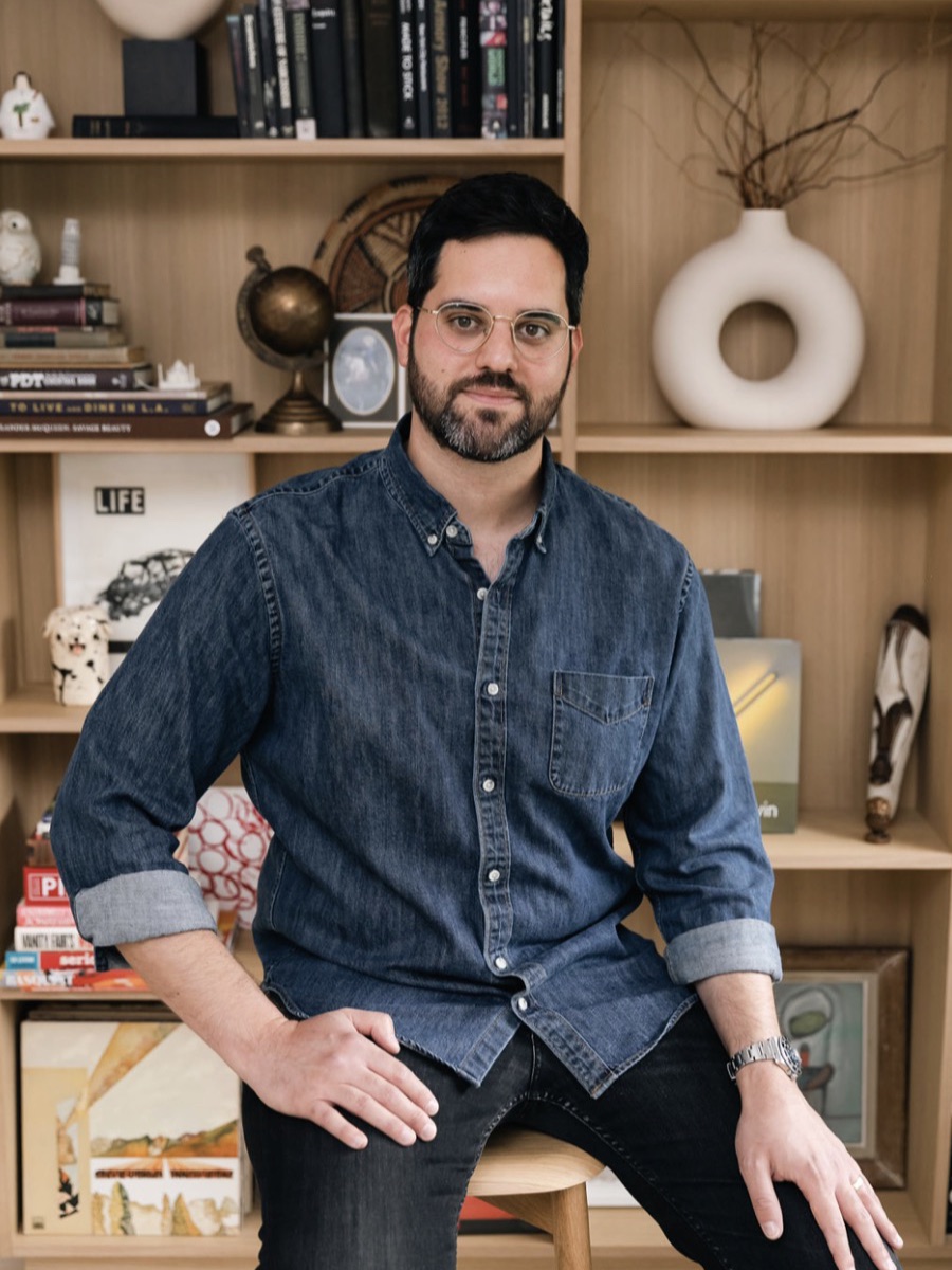 Portrait of a man in a denim shirt leaning on a stool, shelves of books and objects behind him
