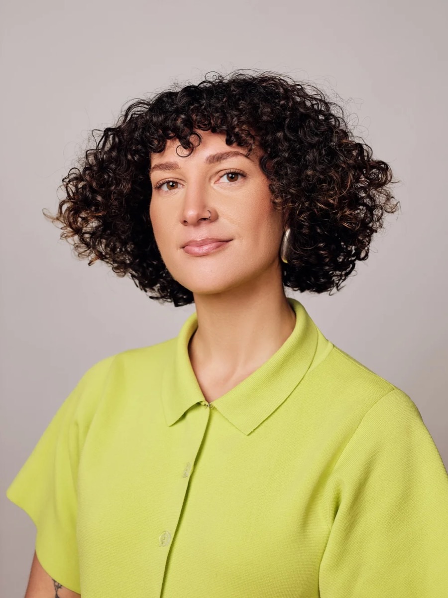 Portrait of a woman with curly dark hair in a lime green collared shirt, against a pale backdrop