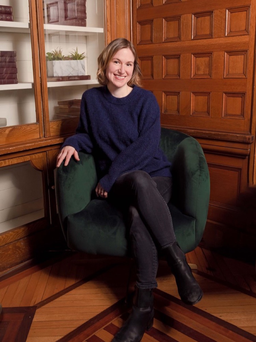 Portrait of a woman in a navy jumper seated in a green armchair, in a wood-panelled room