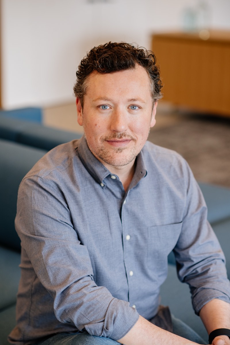Portrait of a man with curly hair in a blue shirt, seated in an office lounge
